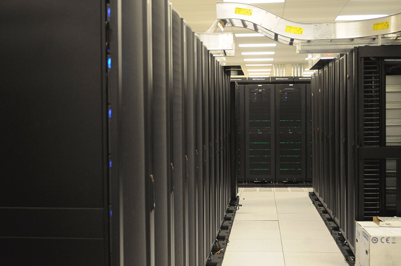 Archive photo of an aisle of black server racks with overhead cabling, a white floor and blue and green status lights.